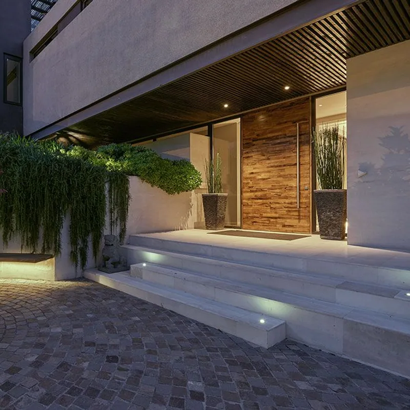 Acceso peatonal iluminado con puerta de madera y alero contemporaneo